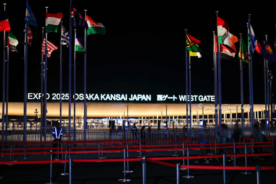 夜の大阪関西万博西ゲート。各国の国旗がはためいている。